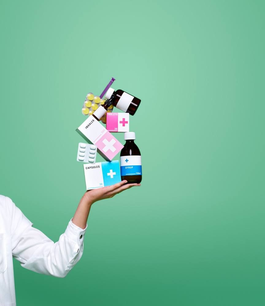 A hand balancing a stack of medicine boxes, bottles and blister packs against a green background